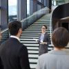 Smiling confident business forum guide with badge on neck standing on stairs and holding clipboards with files while giving tour to participants
